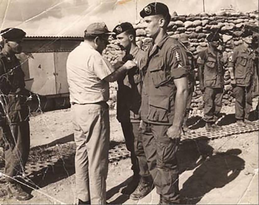 Two men in military uniforms stand at attention while a third man pins something to one of their uniforms. Other people in military uniforms stand at attention in the background.