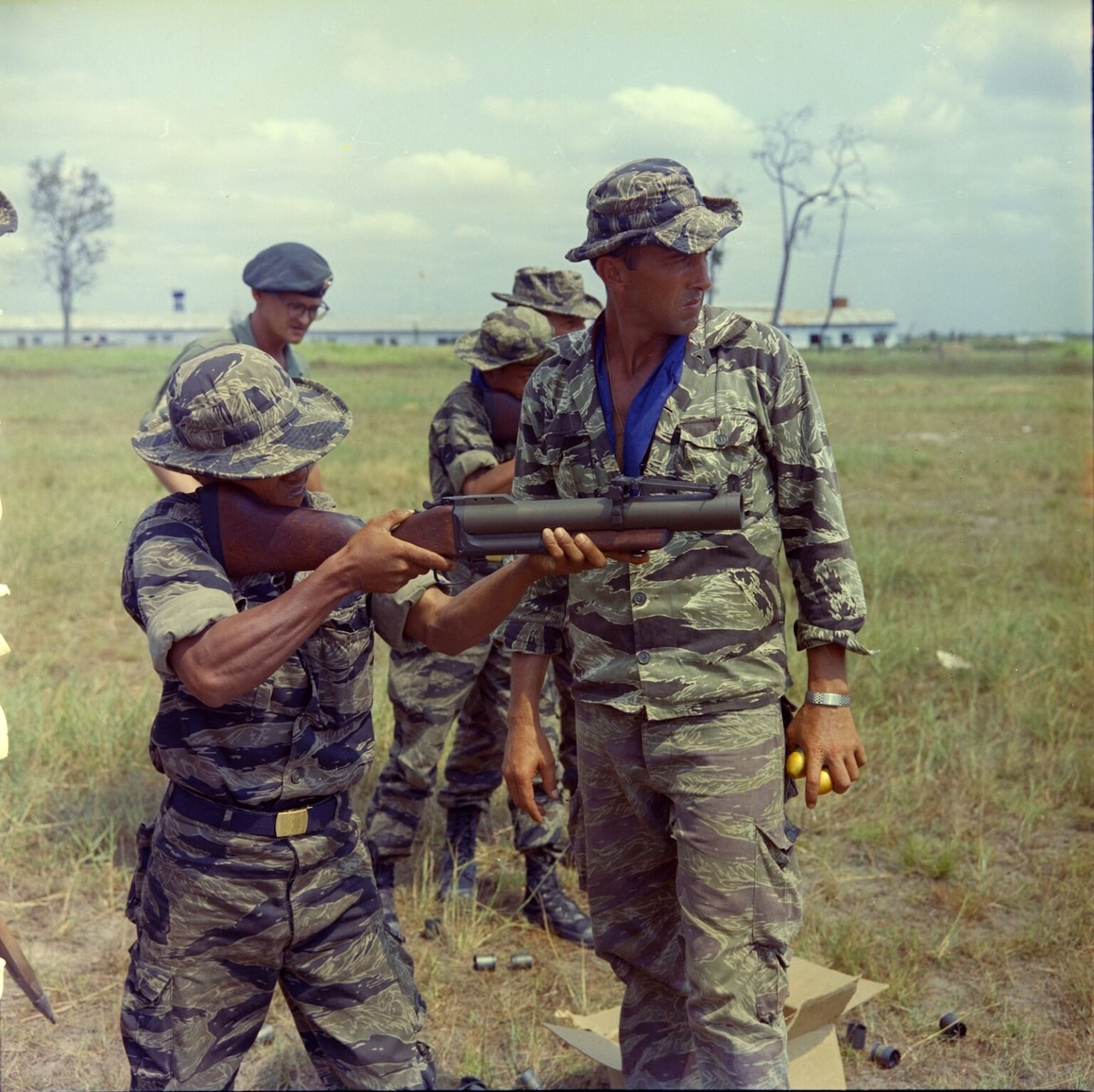 A man in a camouflage military uniform holds a grenade launcher against his shoulder while a man in similar attire beside him looks toward where it’s aimed. Other people in similar attire are standing in the grassy background.