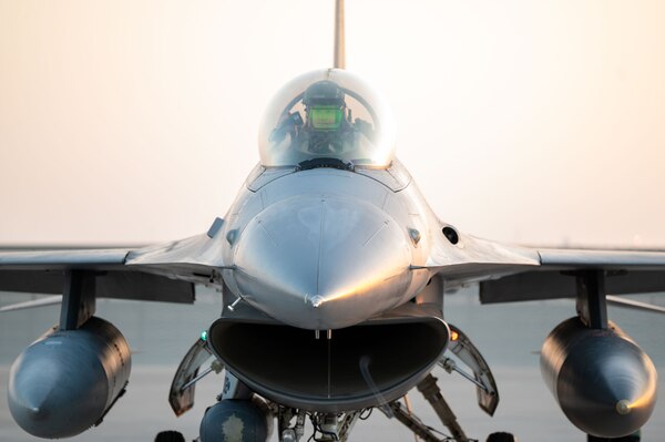 A close-up view of a service member in a flight suit and helmet sitting in the cockpit of a military fighter jet on a tarmac.