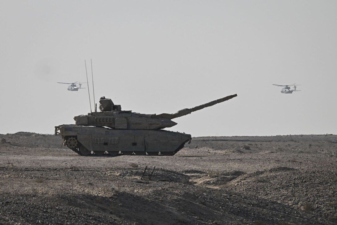 A large military tank points its cannon in the air while sitting in the desert. Two military helicopters are flying in the background.