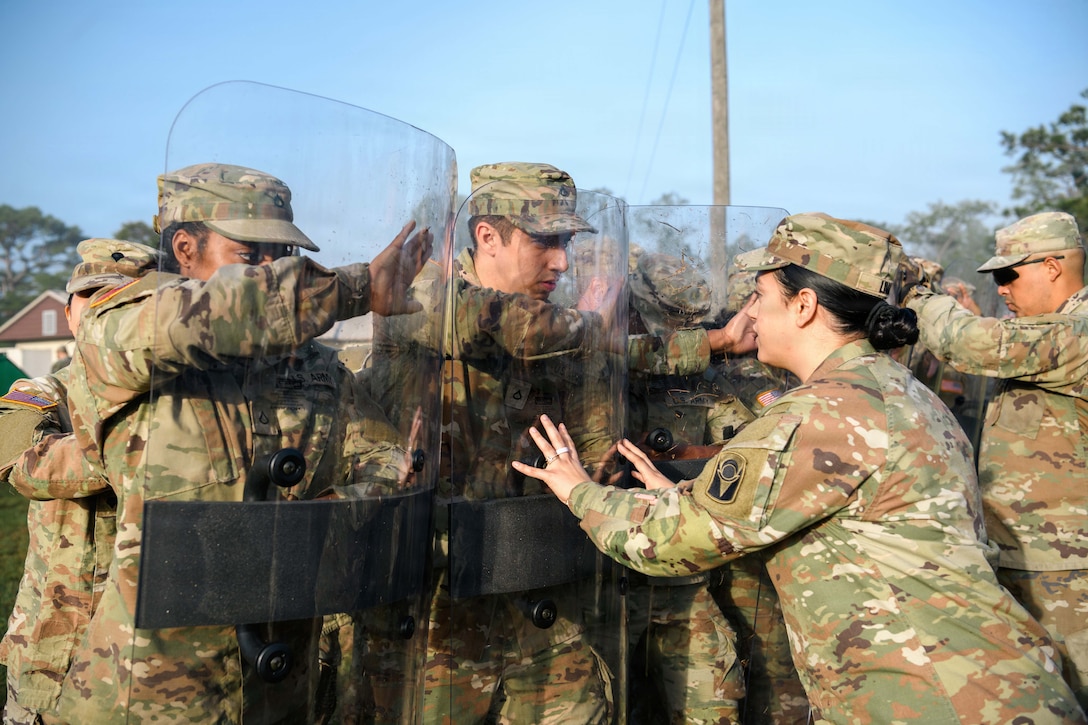 Soldiers stand in a line holding clear shields as a fellow soldier pushes against one under a blue sky.