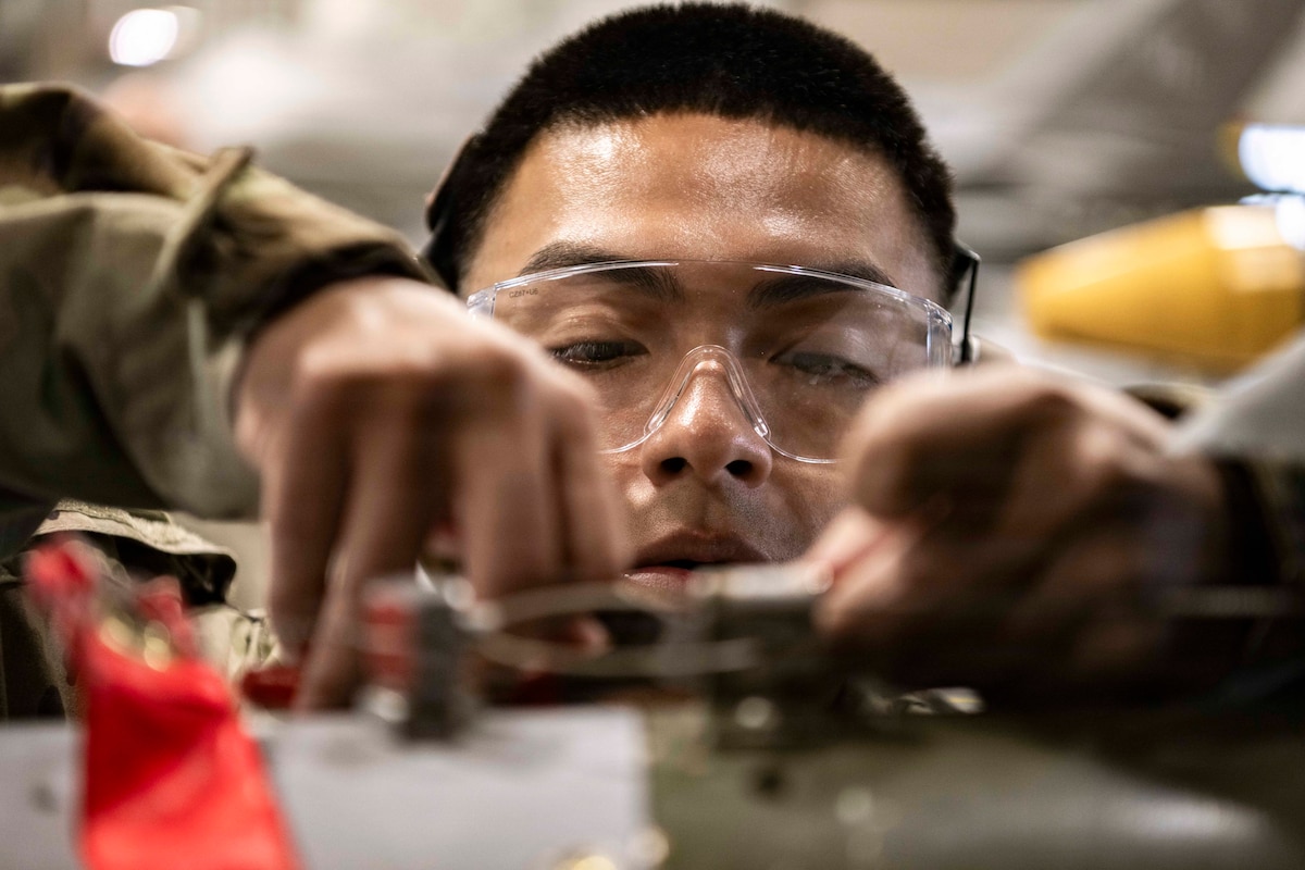 A close-up of an airman in protective eyewear working on a munition in the blurred foreground