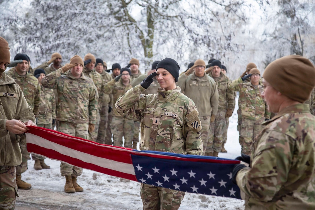 A soldier salutes as two service members hold opposite ends of a folded American flag in front of them while fellow service members salute in formation in the background.