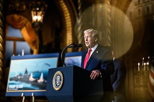 A man speaks from behind a lectern in a room next to a poster of a military vessel.