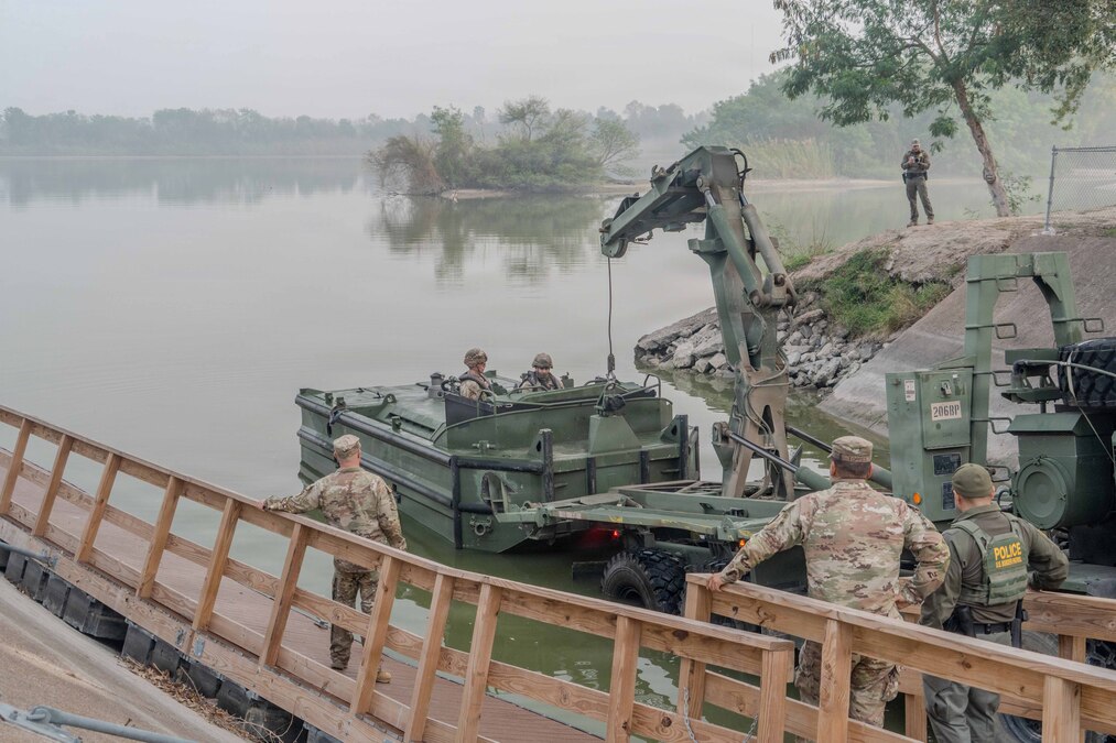 Soldiers stand on a wooden dock and watch as fellow soldiers are lowered into a body of water in a boat on a foggy day.