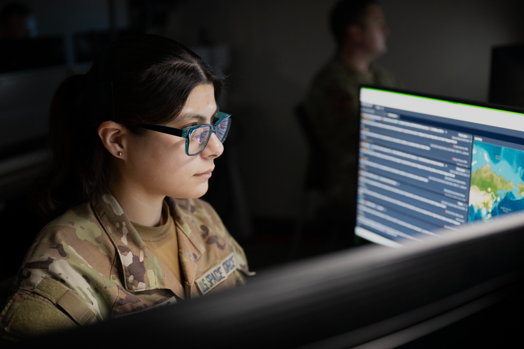 A military woman looks at two unseen screens as her face is illuminated by them in a dimly lit room