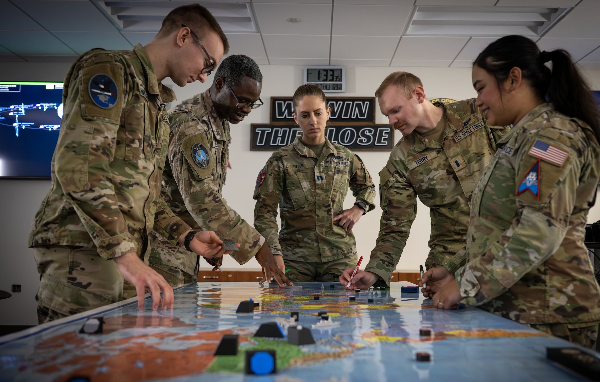 Five military members gather around a wargaming table with a world map and pieces to represent simulated assets