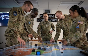 Five military members gather around a wargaming table with a world map and pieces to represent simulated assets