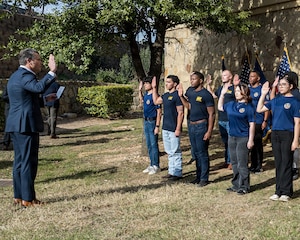 Secretary of War Pete Hegseth stands across from a group of enlistees outdoors, all with hands raised.