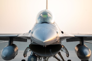 A close-up view of a service member in a flight suit and helmet sitting in the cockpit of a military fighter jet on a tarmac.