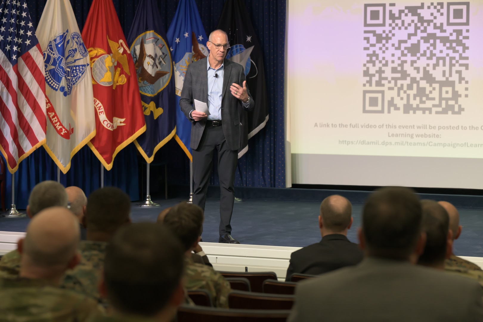 A man in a blazer stands on a stage addressing an audience. U.S. national and military service flags are behind him. The audience is seen from behind, listening to the speaker.