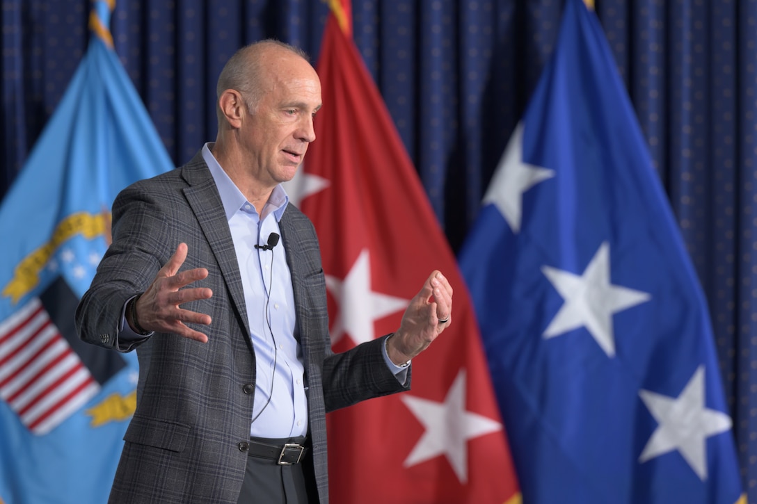A man in a suit jacket speaks and gestures with his hands in front of several military flags. He is wearing a lapel microphone.