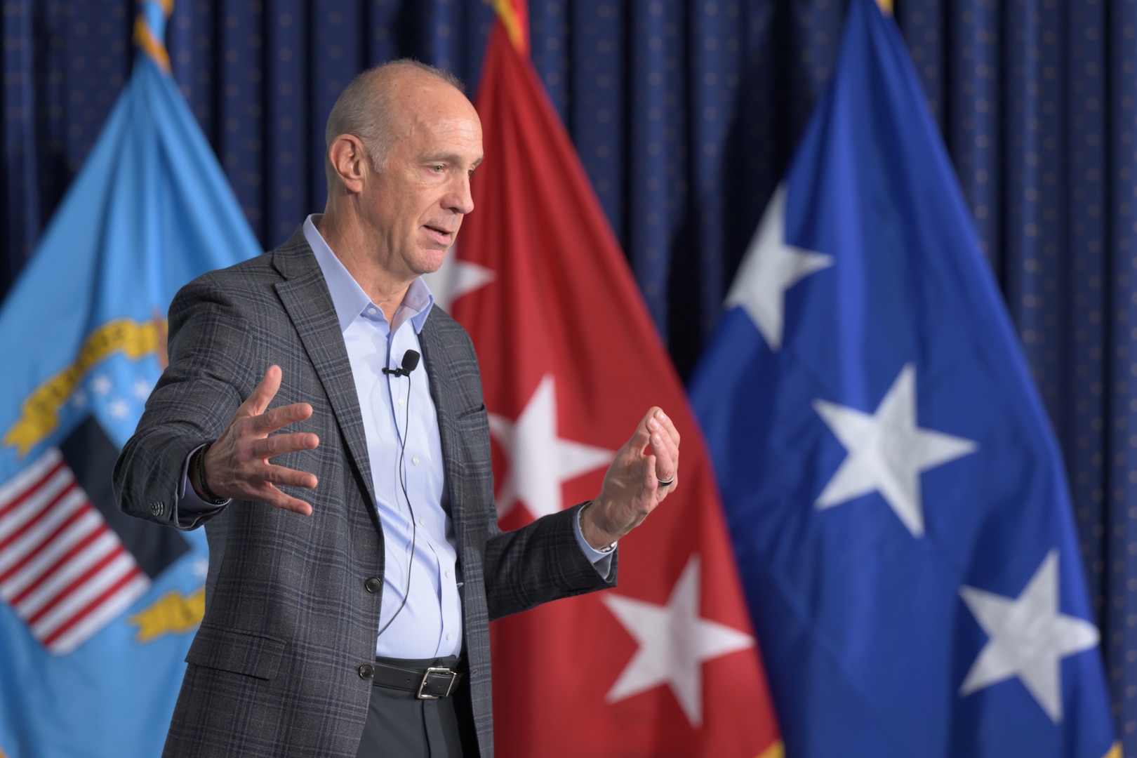 A man in a suit jacket speaks and gestures with his hands in front of several military flags. He is wearing a lapel microphone.