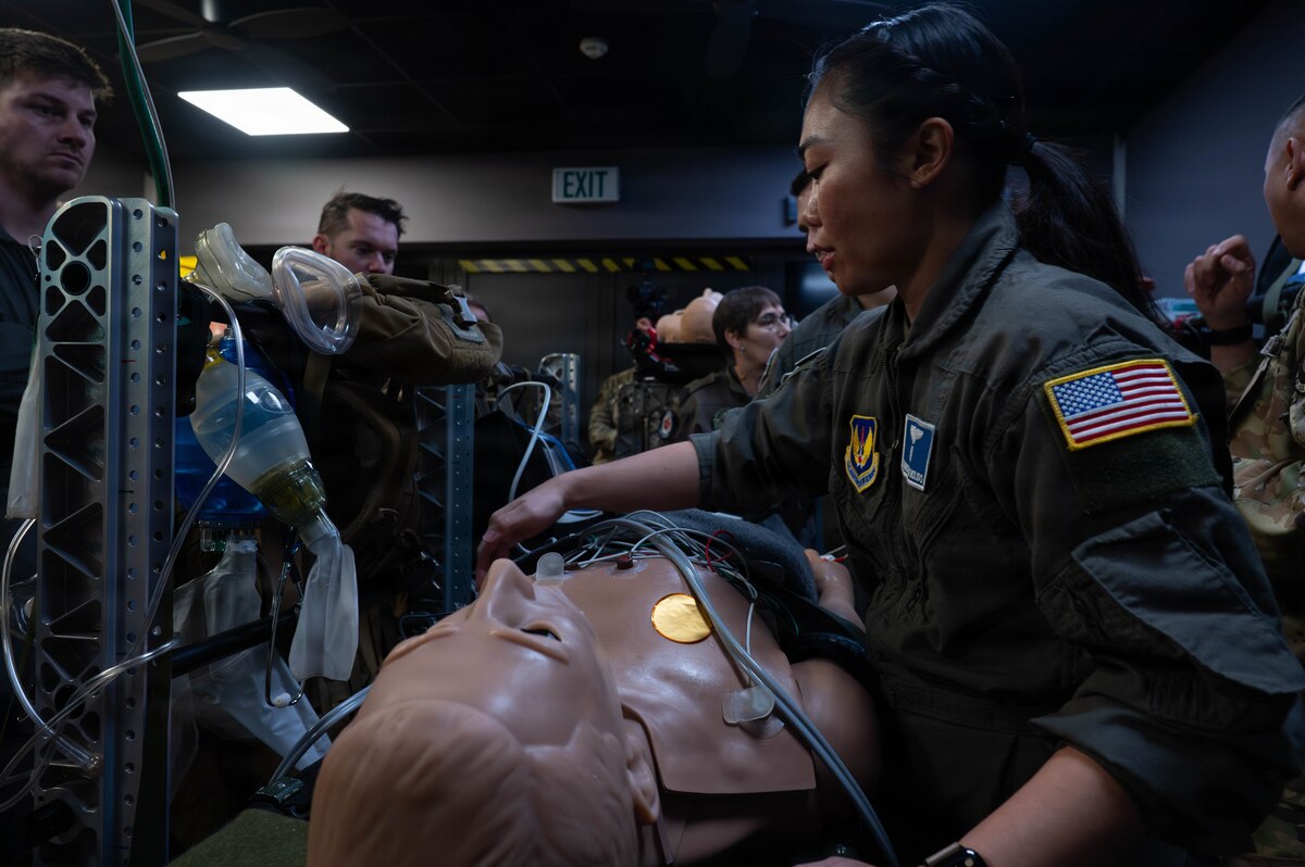 A woman in a military flight suit checks medical equipment attached to a a mannequin on a stretcher. There are several other service members in similar attire standing around her.