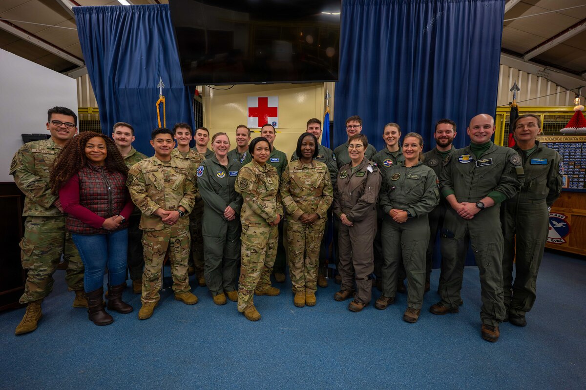 Two dozen service members in camouflage military uniforms and flight suits, and a woman in casual attire stand in two rows as they pose for a photo. There are two large blue curtains hanging in the background and a red cross on the back wall.