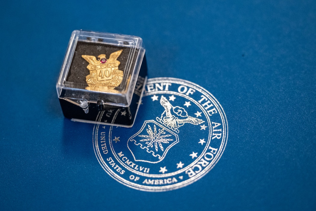 A gold pin, in a clear plastic case, sits on a field of blue with a silver U.S. Air Force seal.