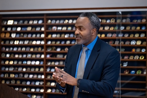 A man in a dark suit stands in front of a case of military challenge coins