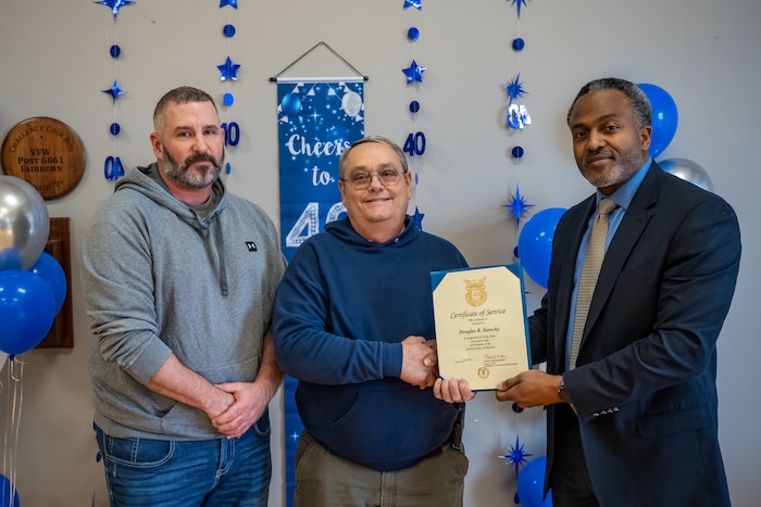 Three men pose for a photo, the man in the center receives a certificate from the man on the right