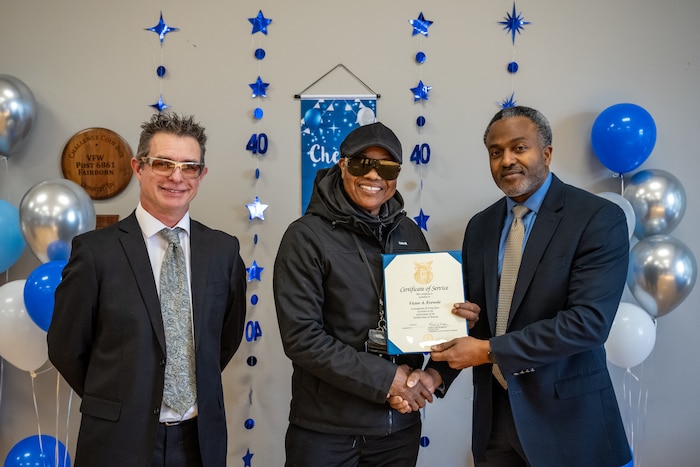 Three men pose for a photo, the man in the center receives a certificate from the man on the right