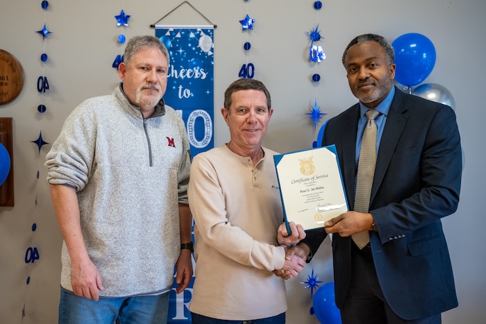 Three men pose for a photo, the man in the center receives a certificate from the man on the right