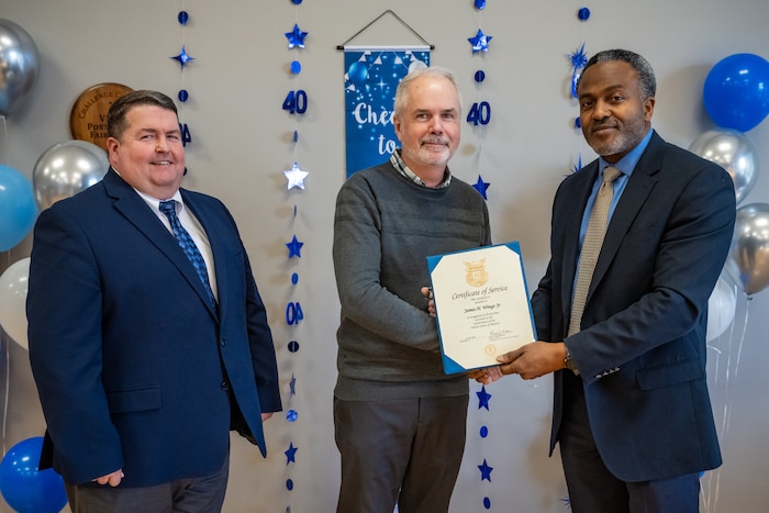Three men pose for a photo, the man in the center receives a certificate from the man on the right
