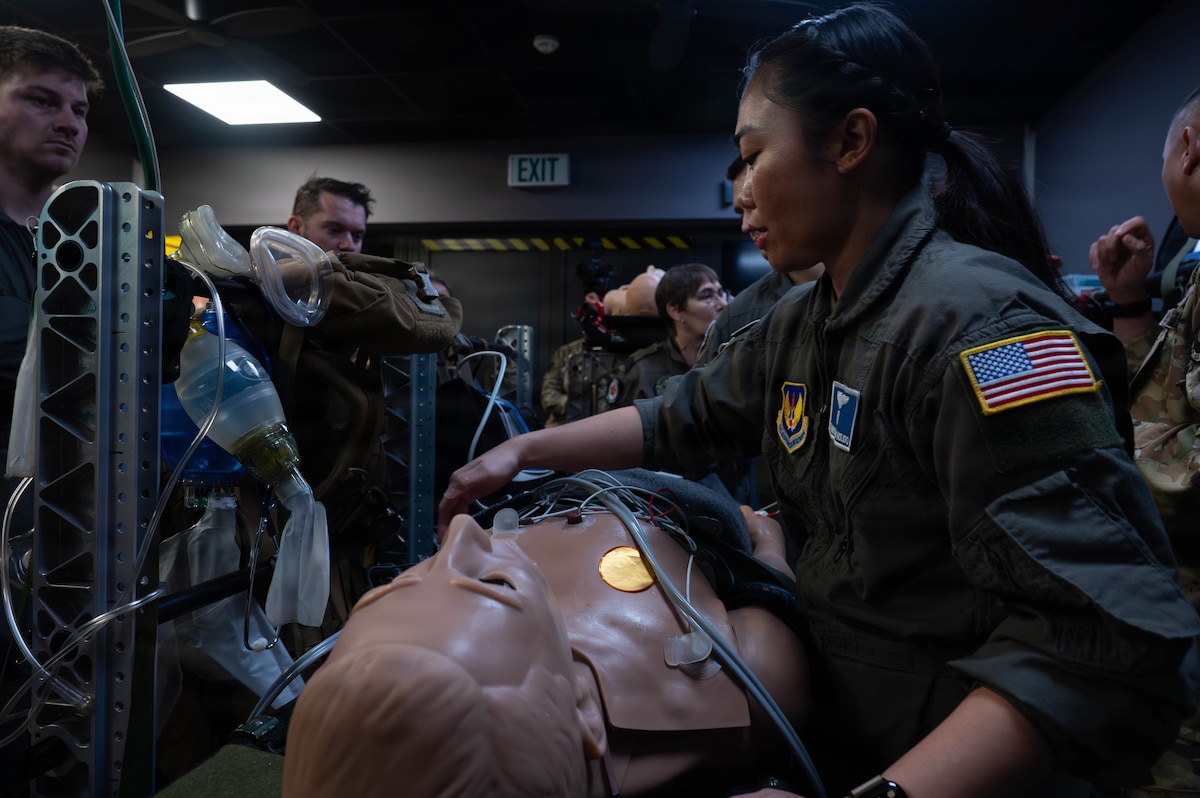 A woman in a military flight suit checks medical equipment attached to a a mannequin on a stretcher. There are several other service members in similar attire standing around her.