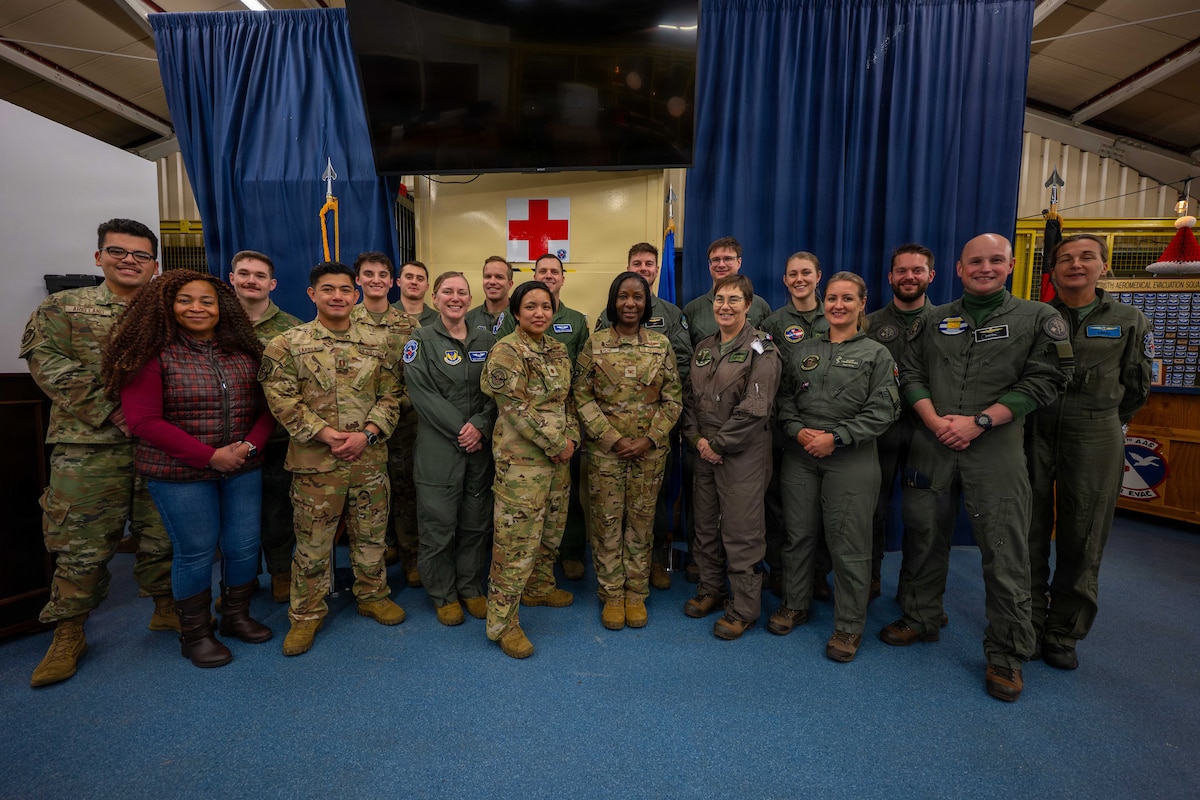 Two dozen service members in camouflage military uniforms and flight suits, and a woman in casual attire stand in two rows as they pose for a photo. There are two large blue curtains hanging in the background and a red cross on the back wall.