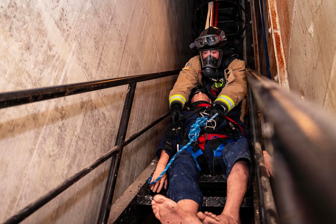 A firefighter wearing full protective gear pulls a mannequin up a small, confined stairwell.
