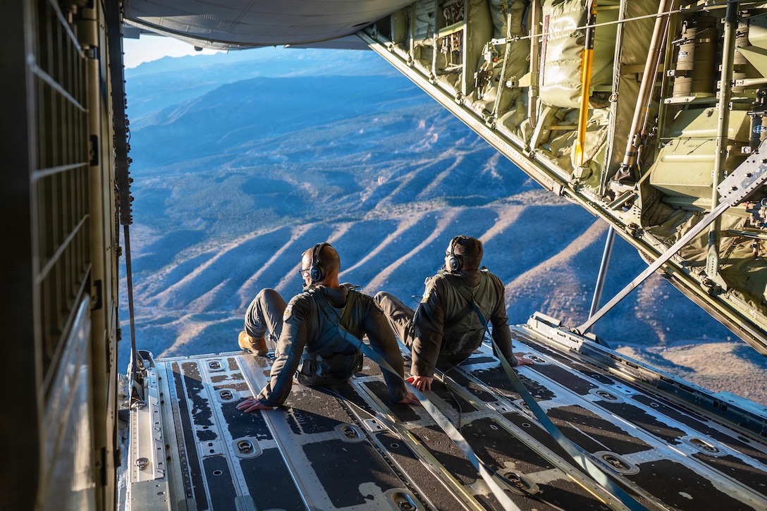 Two people in military uniforms and harnesses sit on the edge of an open military aircraft while flying over mountains.