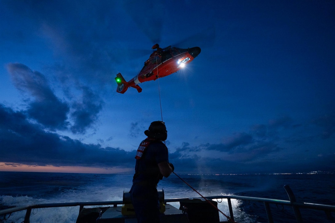 A person holds a tether attached to a military helicopter hovering midair with a spotlight shining downward on the water at night.