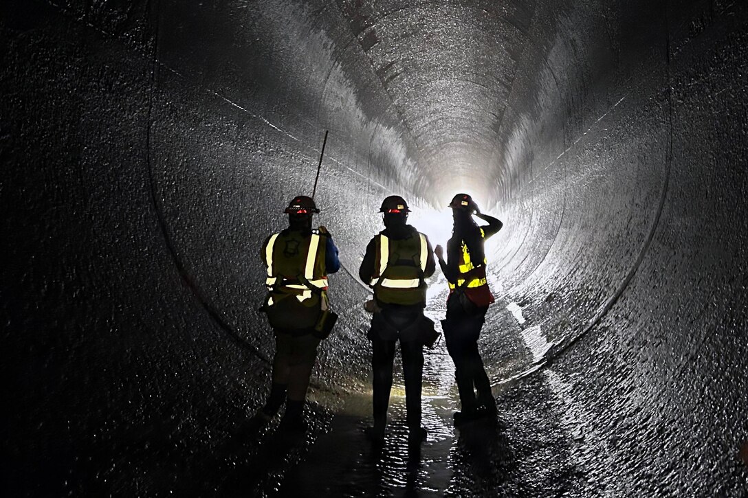 Three people wearing headgear, safety vests and equipment walk through a dark tunnel; a bright light is seen at the other end.