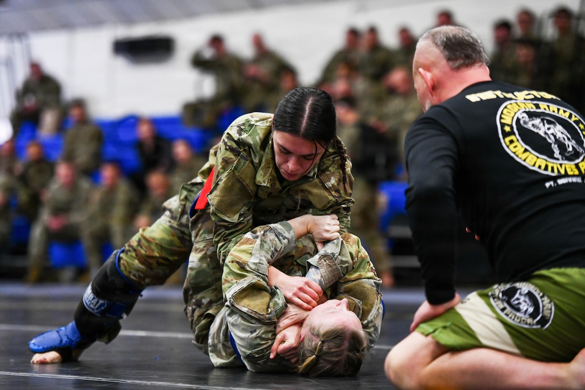 Two women in camouflage military uniforms and protective footgear wrestle on a gym floor in front of a man wearing athletic attire, while others in camouflage military uniforms watch in the background.