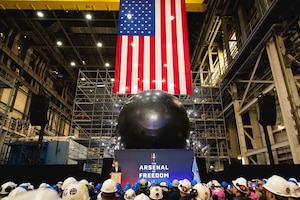 A person standing behind a lectern speaks to a group of people wearing hard hats indoors, with a submarine and American flag in the background.