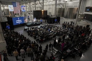 A view from above as the secretary of war stands in front of a large American flag while speaking at a podium to an audience indoors.