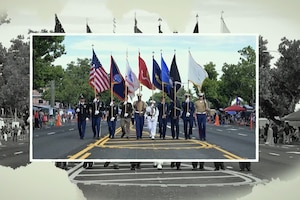 Service members from multiple military branches hold up flags while walking in a row on a street with onlookers watching from both sides.