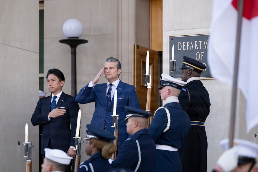 The U.S. secretary of war salutes as the Japanese defense minister puts their hand on their heart on the steps of the Pentagon near service members in ceremonial dress.