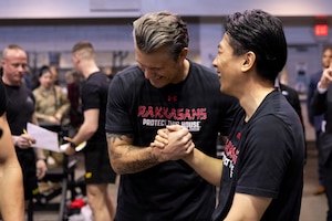 Two men wearing T-shirts smile while clasping each other's right hands. The shirts read "Rakkasans protect the house."