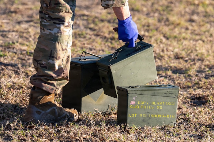 Three green ammo cans sit in the grass, the middle one is being picked up.