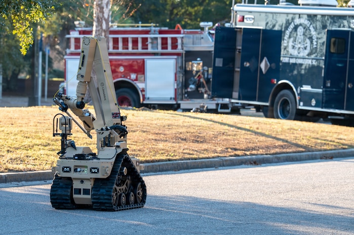 A robot drives in the road toward a firetruck and a large blue truck.