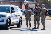 Three Airmen stand together next to a police car. One on the phone, one looking off to the side and one looking at a map.