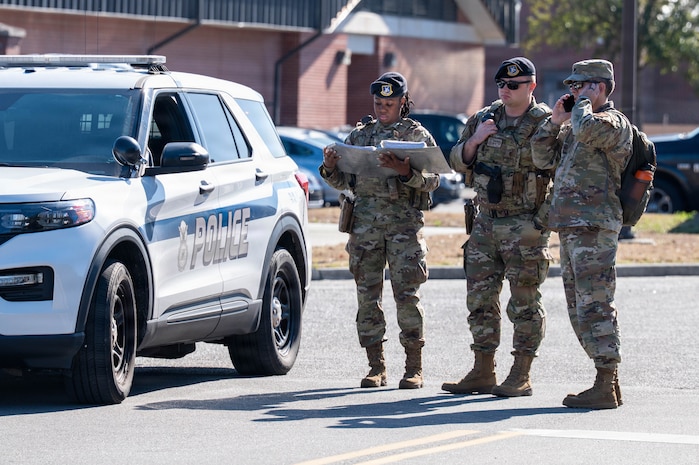 Three Airmen stand together next to a police car. One on the phone, one looking off to the side and one looking at a map.