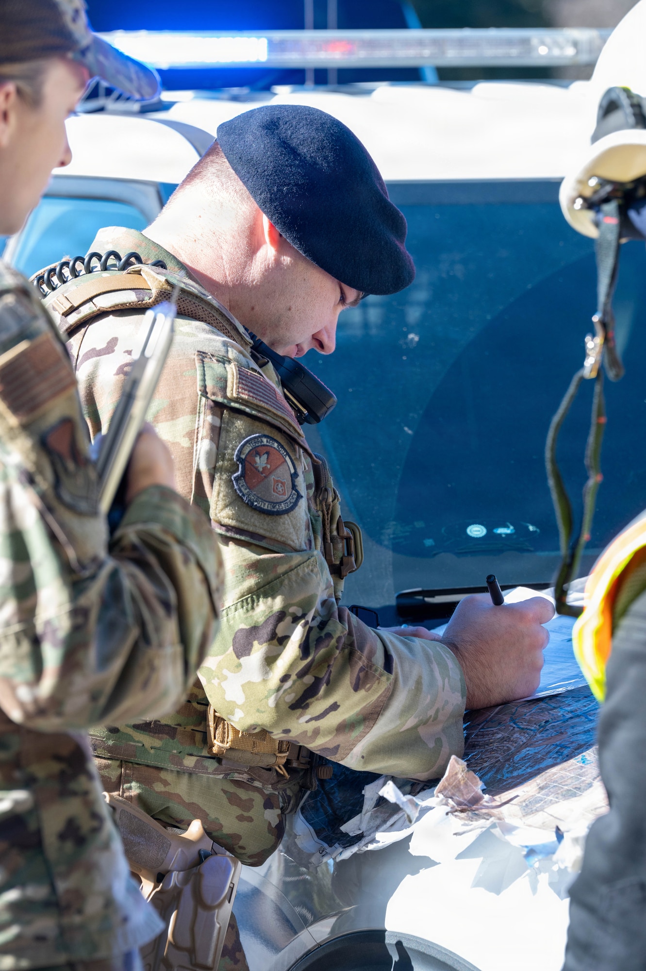 An Airman looks down at a map  while he writes on it on the hood of a car with police lights above him.