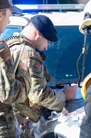 An Airman looks down at a map  while he writes on it on the hood of a car with police lights above him.