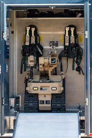 A compact tan robot sits behind an open doorway with equipment hanging behind it
