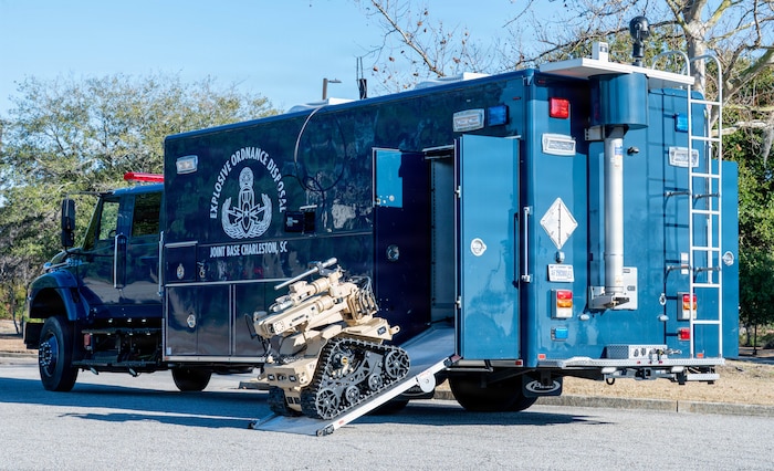 A compact tan robot rolls down a ramp coming out of the side of a large blue truck with the words "Explosive Ordnance Disposal Joint Base Charleston, SC" on the side.