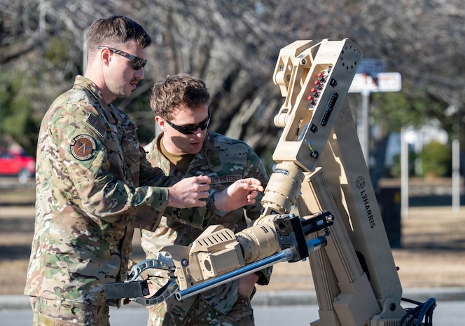 Two Airmen look at the arm of a robot, one reaches for it.