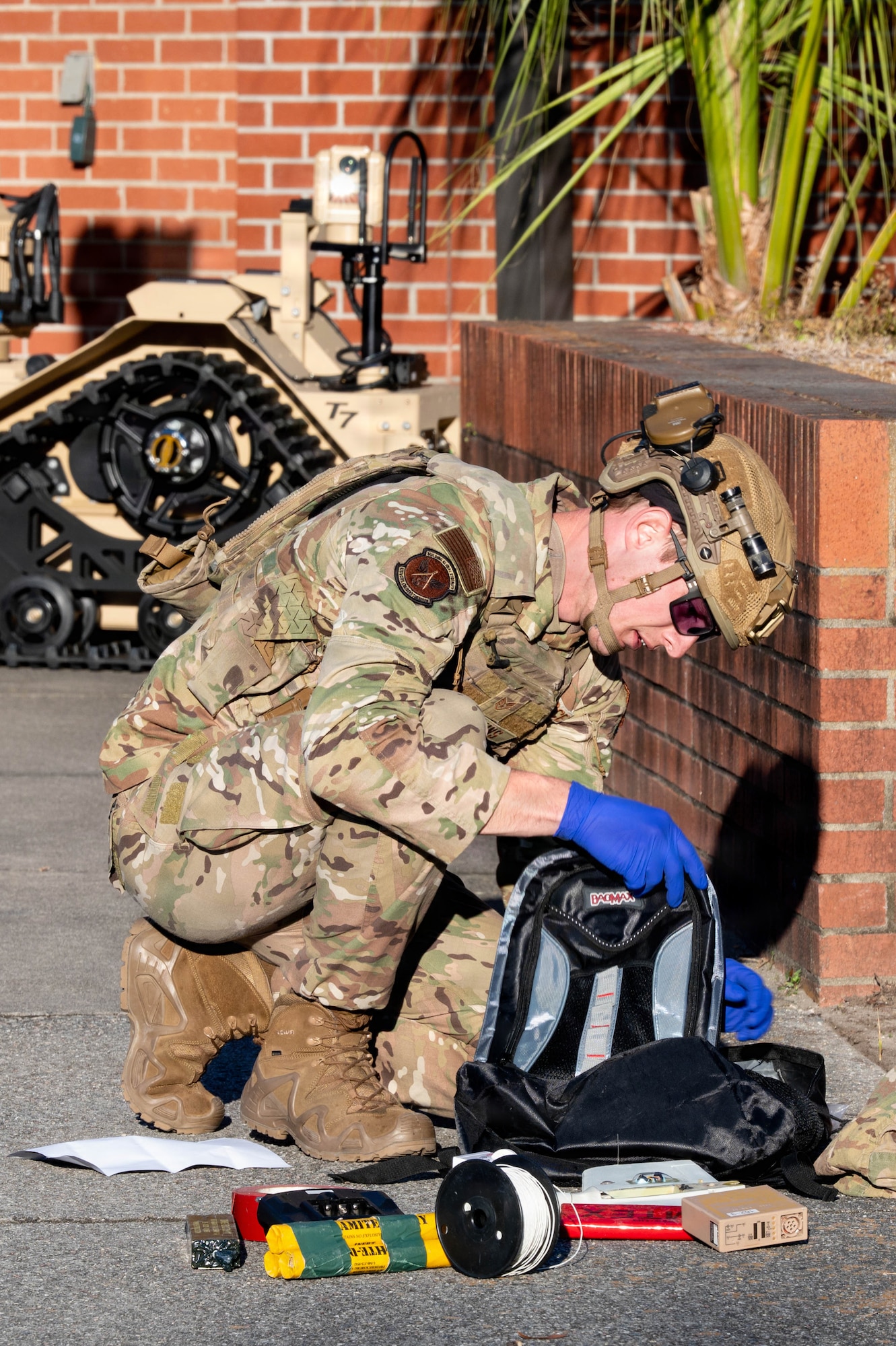 An Airman with a helmet and vest on examines the contents of a backpack with items all around him.