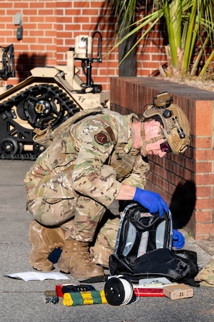 An Airman with a helmet and vest on examines the contents of a backpack with items all around him.