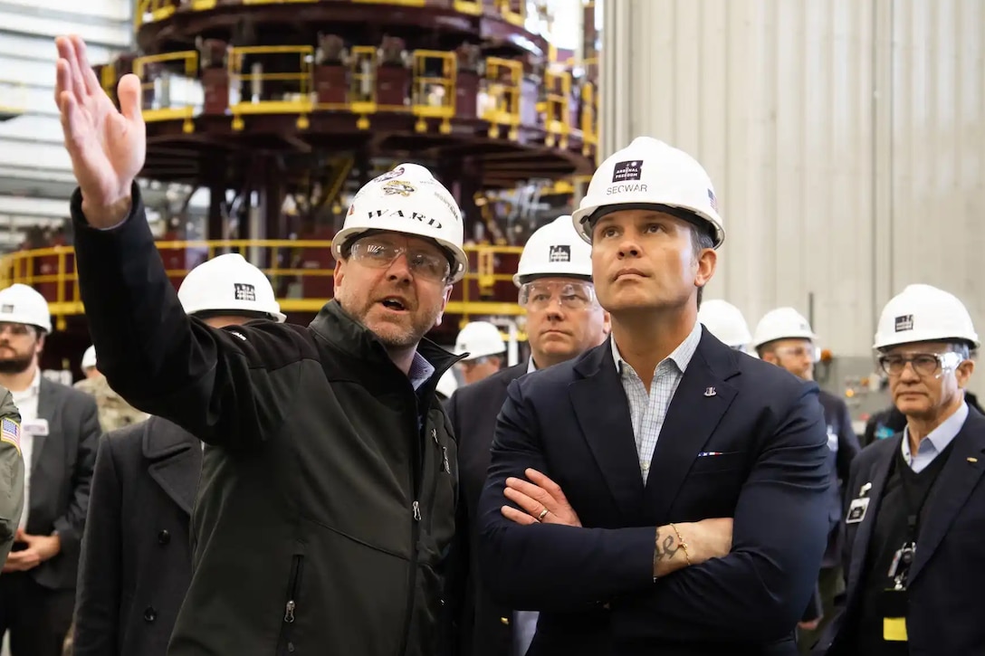 Two people wearing hard hats, one gesturing with a hand, look up inside a factory-like setting, as other people, also in hard hats, stand in the background.