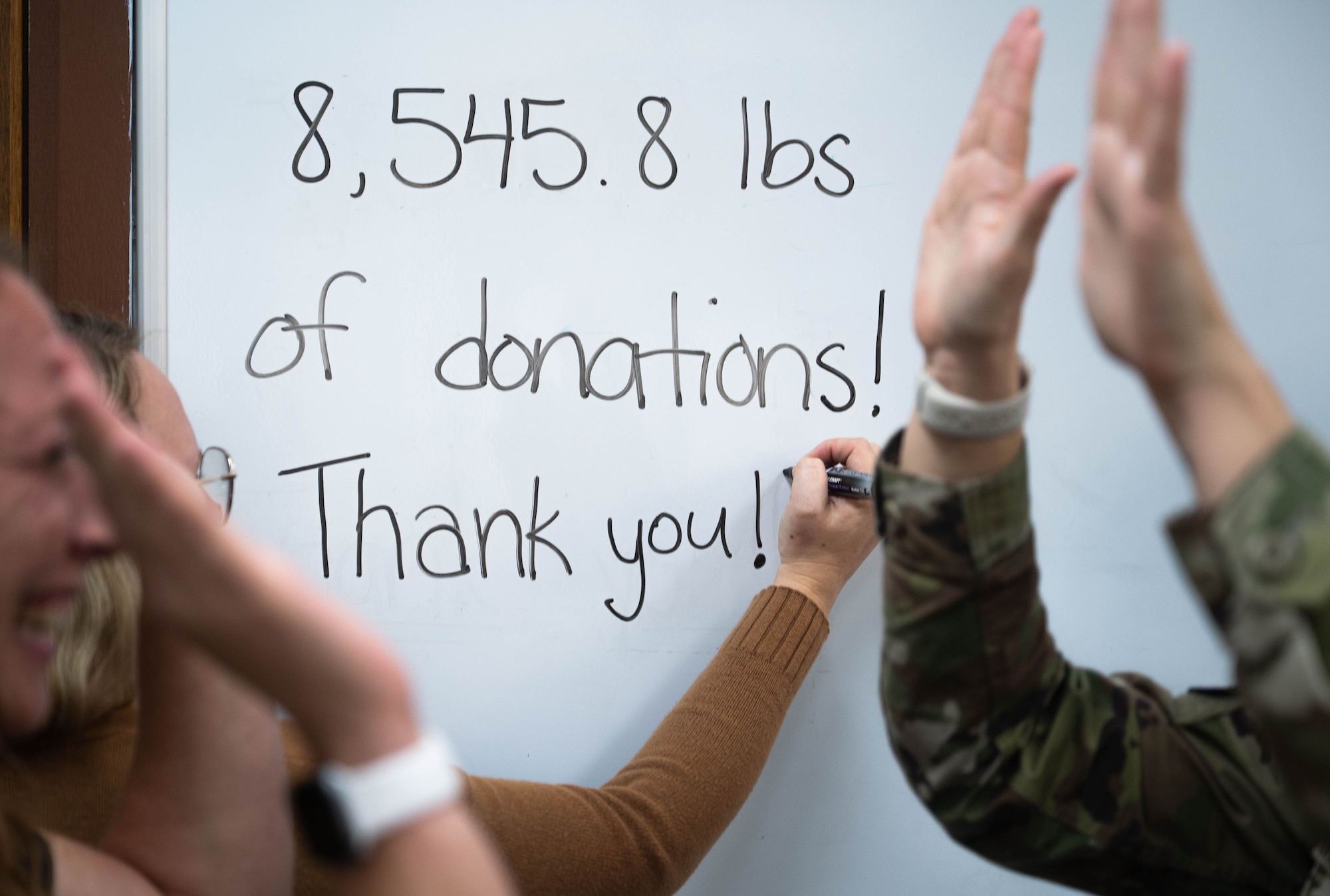 Writing on a white board says "8,545.8 lbs of donations! Thank you!" while people clap and cheer in front of it